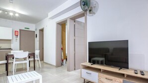 A modern living room with a TV, wooden entertainment unit, and a dining area with a white table and chairs. - Live Where Malta Comes Alive, 2BR in Paceville! (St. Julian`s)