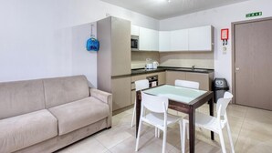 A compact living space with a sofa, a glass-top dining table with chairs, a kitchenette, and a door with an exit sign. - Live Where Malta Comes Alive, 2BR in Paceville! (St. Julian`s)