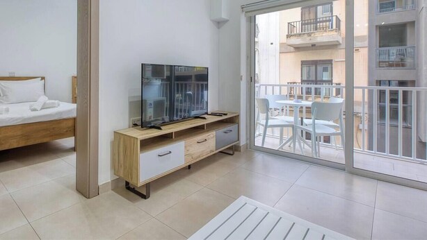 A modern bedroom with a bed, a TV on a wooden stand, a balcony with a table and chairs, and a view of the building's exterior. - Live Where Malta Comes Alive, 2BR in Paceville! (St. Julian`s)