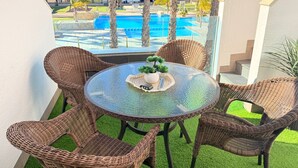 Uma varanda com cadeiras de vime, uma mesa de vidro e vista para uma piscina e edifícios modernos.