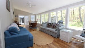 A spacious living room with a blue sofa, a patterned sofa, a dining area with chairs, and large windows offering a view of the outdoors. - Private Orleans Getaway Near Bakers Pond & Beaches (2422) (Orleans)