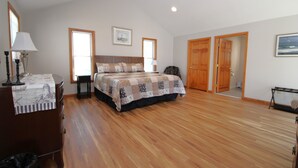 A bedroom with a wooden floor, a bed with a quilt, a nightstand, a lamp, a dresser, a chair, and a door leading to another room. - Private Orleans Getaway Near Bakers Pond & Beaches (2422) (Orleans)
