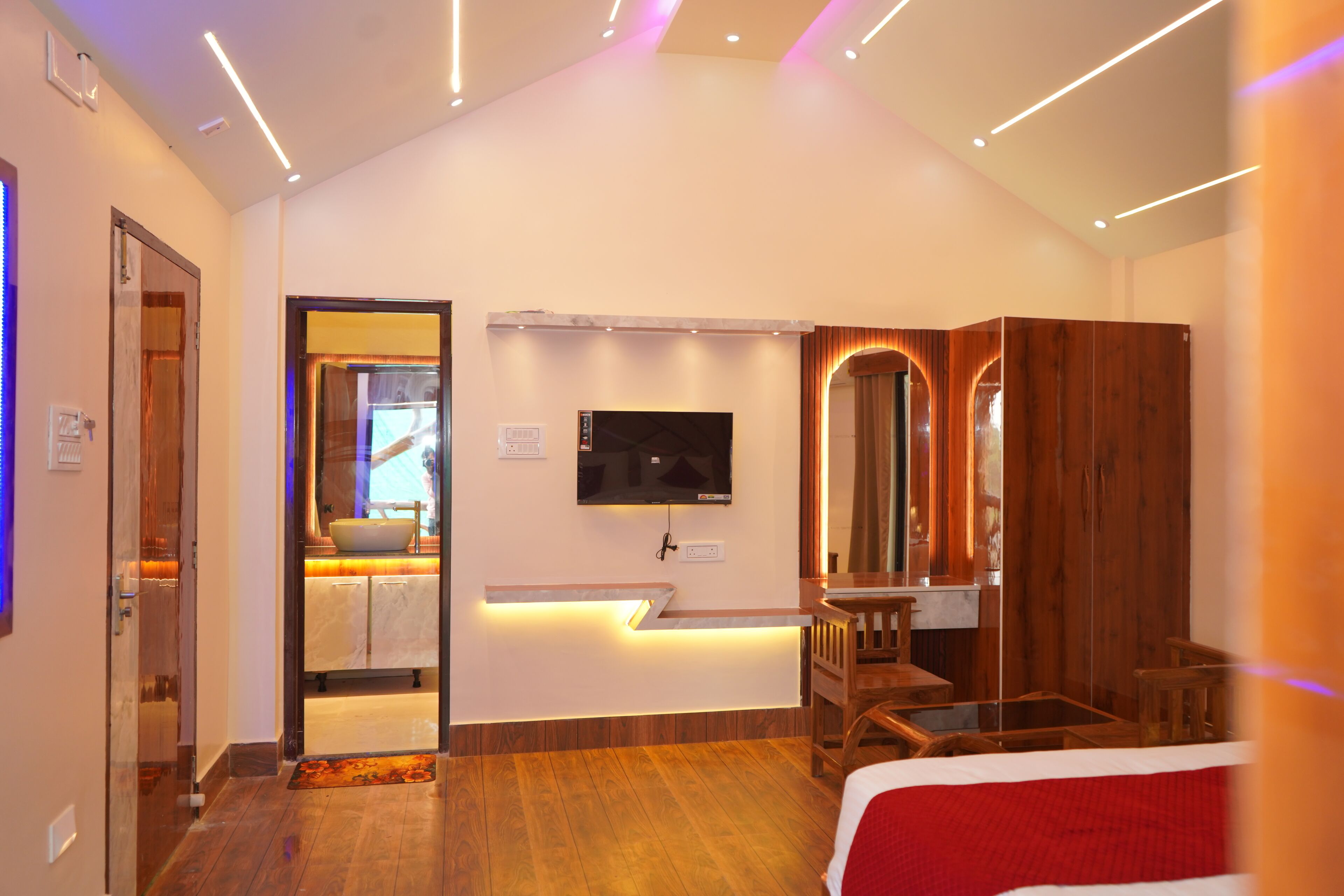 A modern hotel room with a wooden floor, a bed, a desk, a TV, and a bathroom visible through an open door.