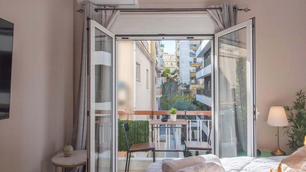 Une chambre avec une grande porte-fenêtre coulissante donnant sur un balcon, un lit avec une literie blanche, une petite table ronde avec une lampe et une plante en pot.