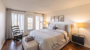 Un dormitorio con una cama grande, una silla, una mesita de noche y un ventanal con cortinas.