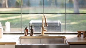 Una cocina con un lavamanos de acero inoxidable, un grifo dorado y vistas a un área verde a través de ventanas amplias.
