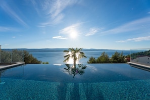 Cielo azzurro intenso con riflessi del sole su una piscina, circondata da alberi e con vista sul mare e sulle montagne lontane.