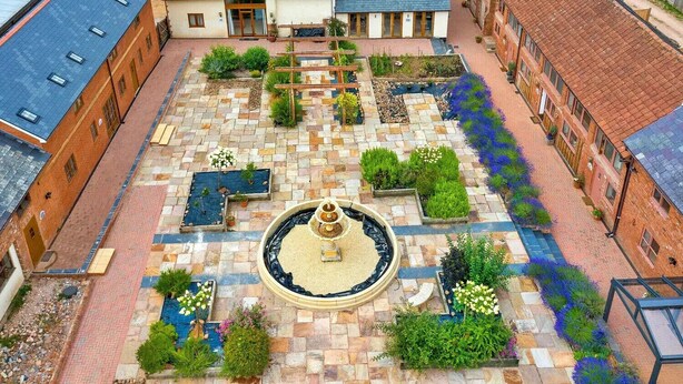 Ein Innenhof mit einem zentralen Brunnen, umgeben von Ziegelgebäuden mit zahlreichen Fenstern und einem Gartenbereich.