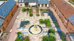 A courtyard with a central fountain, surrounded by brick buildings with multiple windows and a garden area with various plants. - Lime Cottage, Great Houndbeare Farm (Aylesbeare)