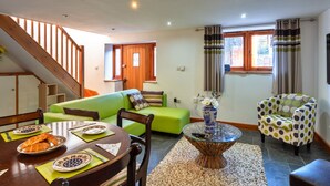 A living room with a green sofa, a dining table with plates of food, a wooden chair, a coffee table with a vase, and a staircase leading to another room. - Lime Cottage, Great Houndbeare Farm (Aylesbeare)
