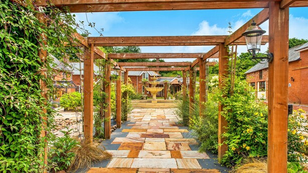 A wooden pergola with a stone pathway leading to a fountain, surrounded by greenery and brick buildings. - Lime Cottage, Great Houndbeare Farm (Aylesbeare)