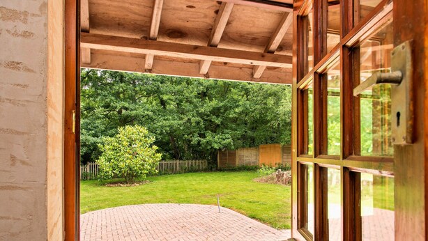 A wooden-framed window with a view of a brick patio and a grassy backyard with trees and a wooden fence.