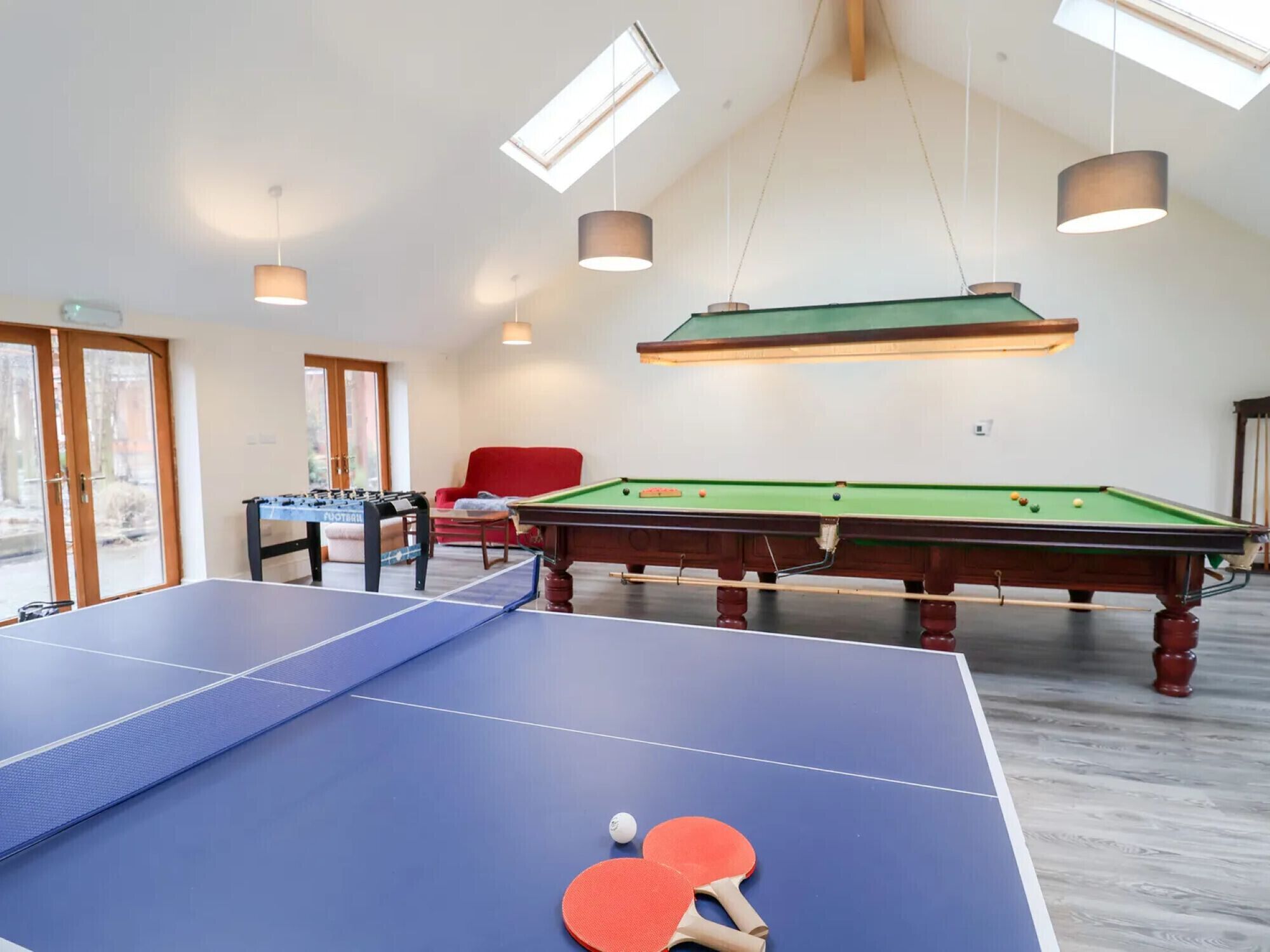 Une pièce équipée d’une table de ping-pong, d’une table de billard et d’un baby-foot. Il y a des fenêtres et un fauteuil rouge.