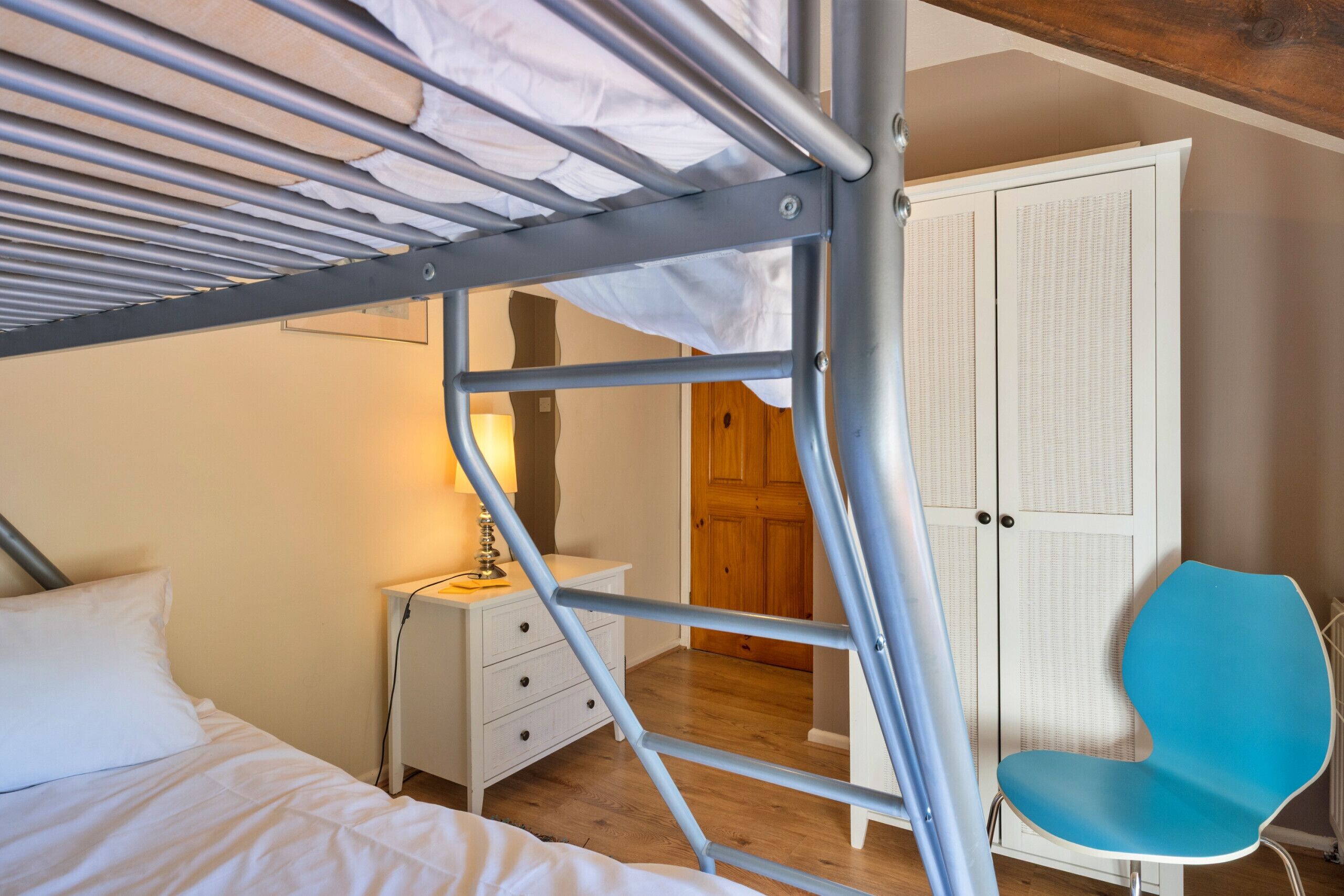 A bunk bed with a metal frame, a white bedside table with drawers, a blue chair, and a wardrobe with louvered doors.
