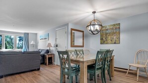 A modern living room with a dining area, wooden flooring, and a large window with curtains. - Perfect Wells Maine getaway, Village by the Sea Condo! (Wells)