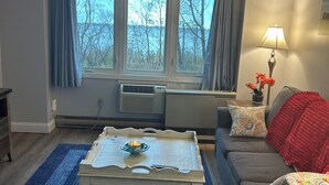 A living room with a sofa, a coffee table, a lamp, and a window with curtains. - Perfect Wells Maine getaway, Village by the Sea Condo! (Wells)