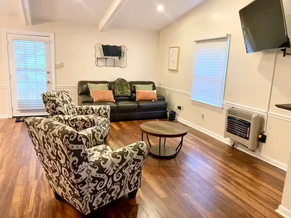 A living room with patterned armchairs, a sofa, a round coffee table, and a flat-screen TV mounted on the wall. - Modern Family Living – Near Food, I-85 & Hospital (Henderson)