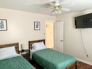 A bedroom with two beds, a ceiling fan, a TV mounted on the wall, and framed artwork on the wall. - Modern Family Living – Near Food, I-85 & Hospital (Henderson)