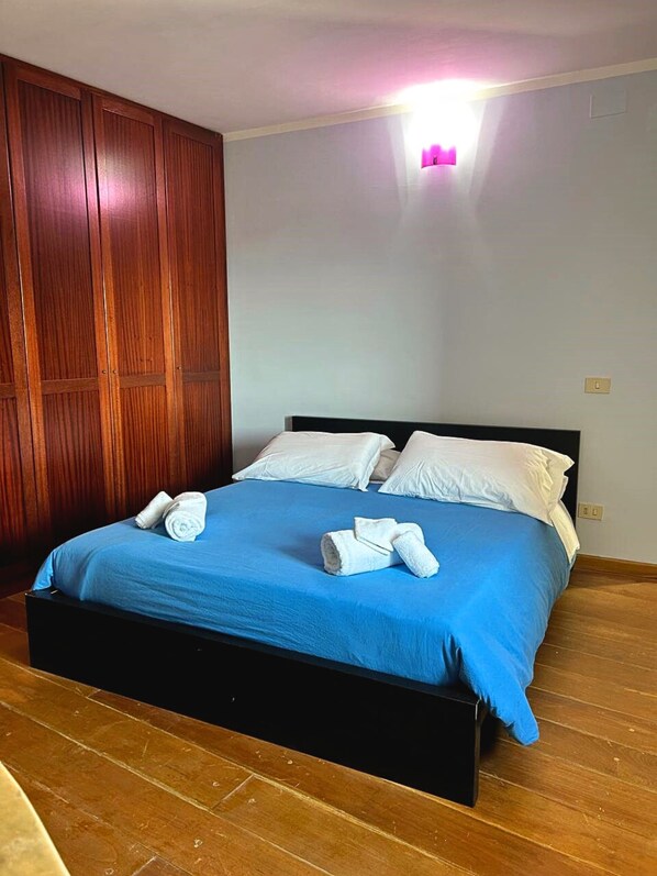 A neatly made bed with blue bedding and white pillows, wooden wardrobe, and a light switch on the wall.