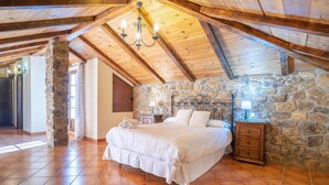 Un dormitorio con pared de piedra, vigas de madera en el techo, una cama y una mesita de noche con lámpara.