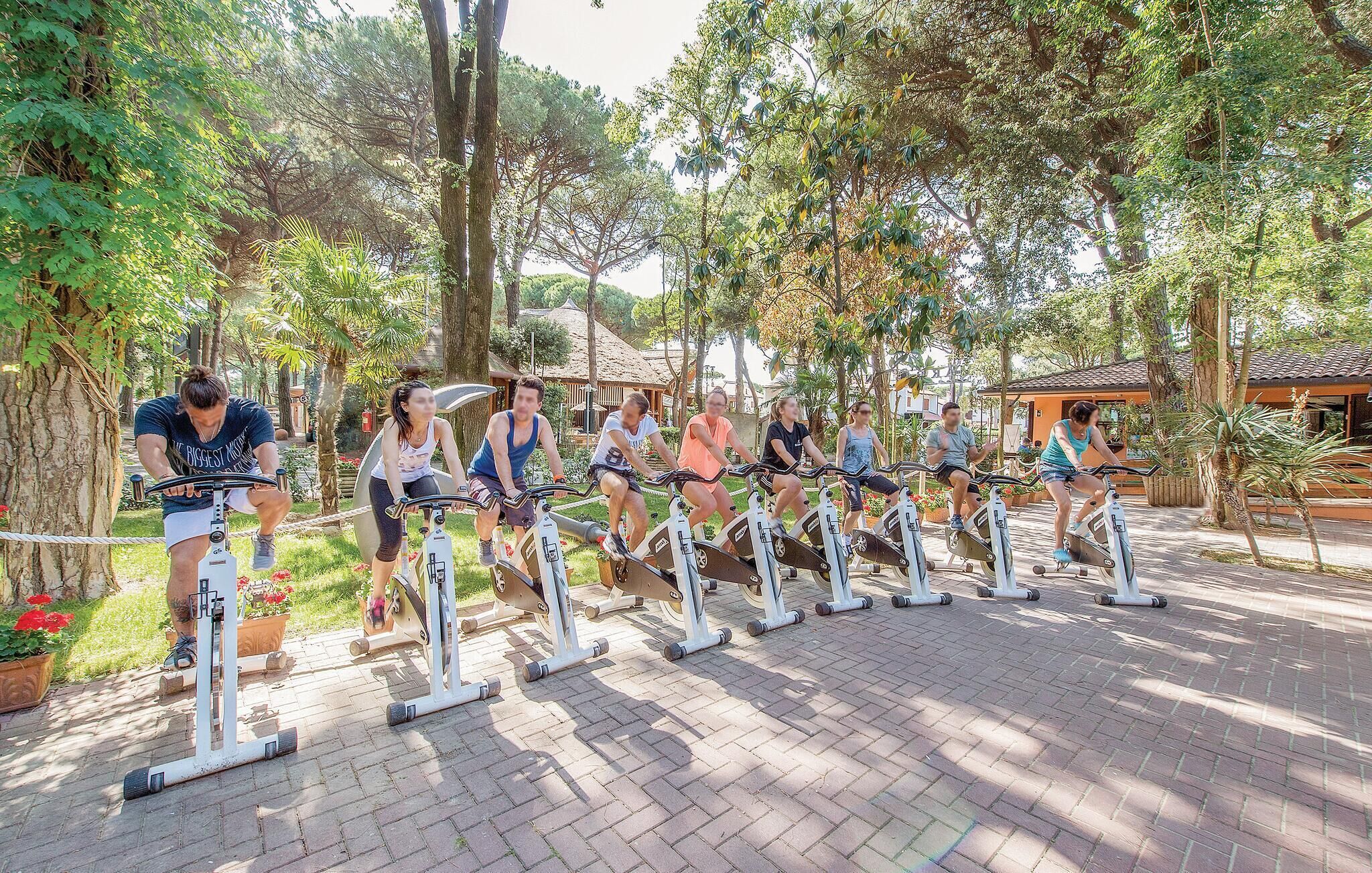 A group of people using stationary bikes outdoors in a park-like setting with trees and a brick pathway.