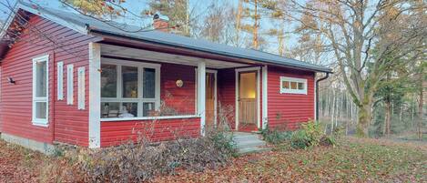 Une maison en bois rouge, dotée d’un porche, entourée d’arbres et de feuilles mortes.