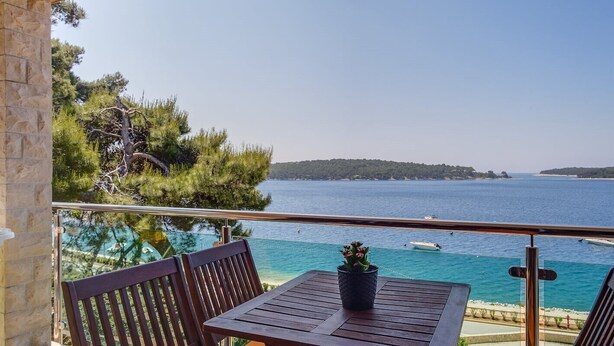 Een balkon met een houten tafel en stoelen, met uitzicht op een heldere blauwe zee met boten en een eiland in de verte.