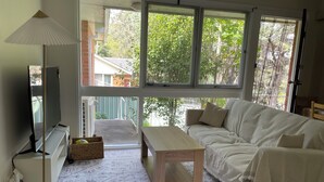 Un salon chaleureux avec un canapé, une petite table en bois, une lampe sur pied et un téléviseur. De grandes fenêtres offrent une vue sur l’extérieur.