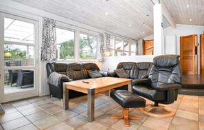 Un salon chaleureux avec un canapé en cuir, une table basse en bois et un fauteuil en cuir noir. De grandes fenêtres, ornées de rideaux, laissent entrer la lumière naturelle et offrent une vue sur l’extérieur.