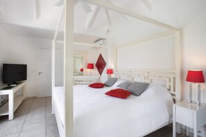 A hotel room with a four-poster bed, a television on a stand, and a desk with a lamp. - Encore   Tropical Gardens and Ocean Views (Saint Martin)