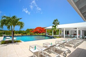 A pool area with lounge chairs and tables, surrounded by palm trees and a red flowering tree. - Encore   Tropical Gardens and Ocean Views (Saint Martin)