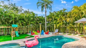 Ein Swimmingpool mit einem rosa Flamingo-Schwimmsack, umgeben von Palmen und einer hölzernen Spielanlage mit Rutsche.