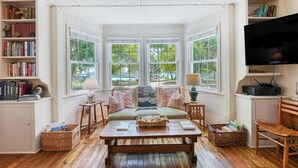 A cozy living room with a sofa, coffee table, and bookshelves, featuring large windows and a television. - 4 Bedroom 2 Bath Inlet Front Home with Deep Water Access (Murrells Inlet)