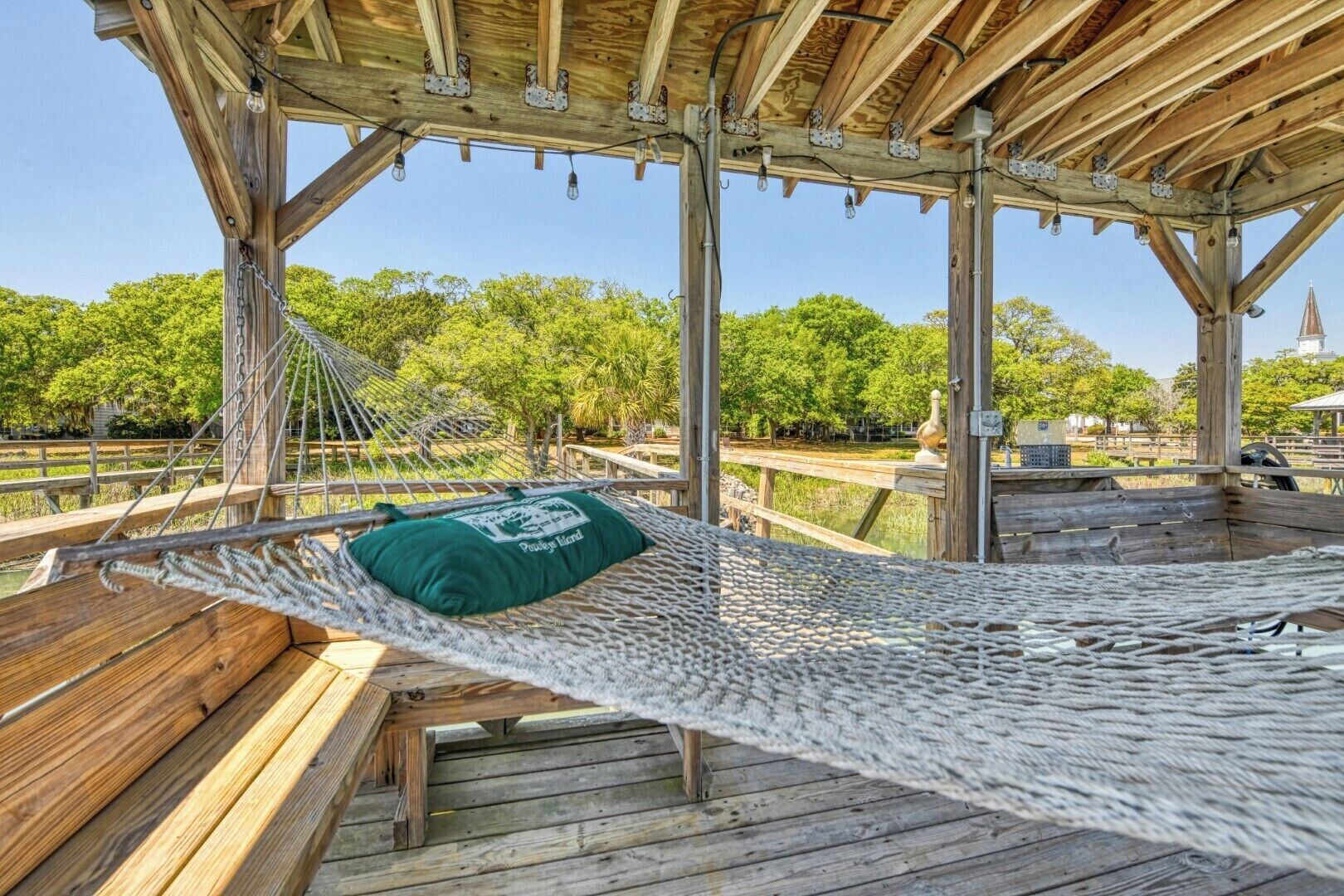 A wooden deck with a hammock and a bench, surrounded by trees and a clear sky.