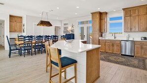 A spacious kitchen with wooden cabinets, a central island, and a dining area with blue chairs. - Coastal Luxury | Beachfront Home | Lagoon Views (Gulf Shores)
