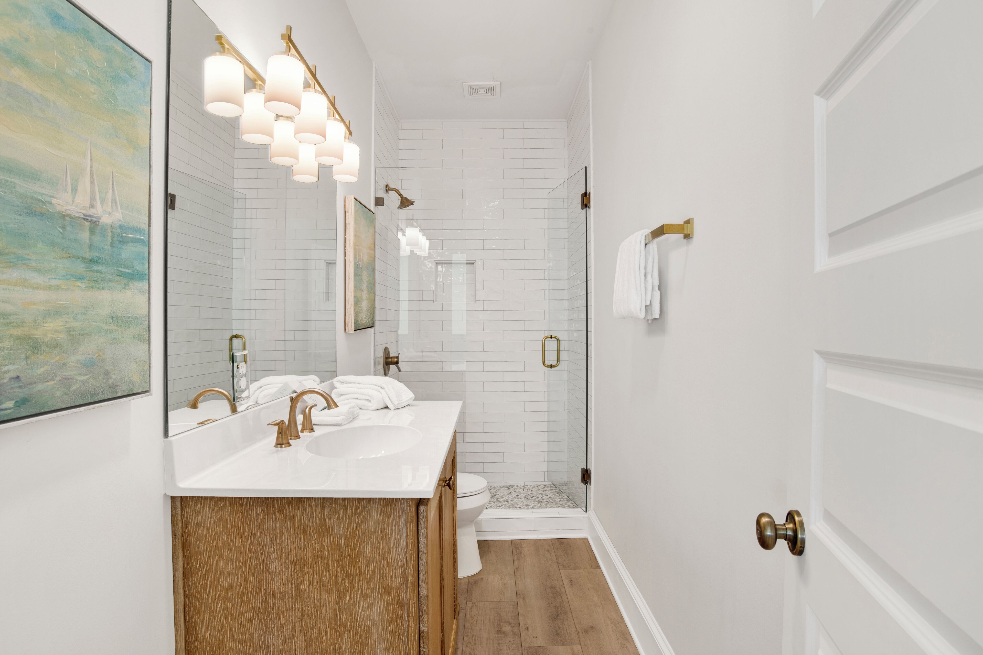A bathroom with white subway tiles, a wooden vanity, a white sink, a gold faucet, and a glass shower enclosure.