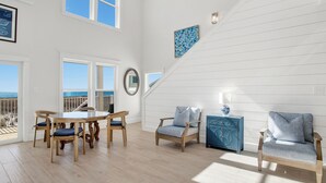 A spacious room with a wooden floor, a dining table with chairs, a blue sofa, and a staircase leading to a balcony with a view. - Coastal Luxury | Beachfront Home | Lagoon Views (Gulf Shores)