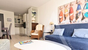 A living room with a white coffee table, a blue sofa, and a wicker chair. There is a kitchen in the background with white cabinets and a refrigerator. - GRAND T2 AVEC TERRASSE, GARAGE ET PISCINE (Sète)
