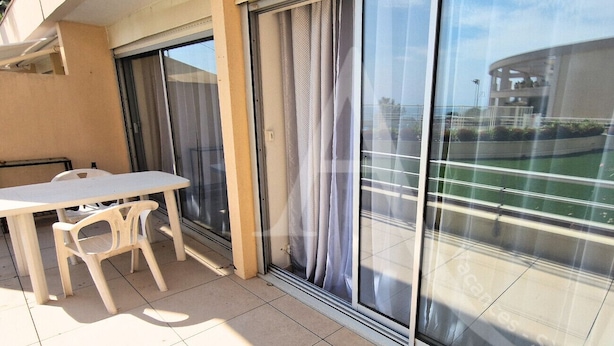 A balcony with a table and chairs, a sliding glass door, and a view of a building across the street. - GRAND T2 AVEC TERRASSE, GARAGE ET PISCINE (Sète)