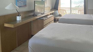 A hotel room with two beds, a desk with a computer, a ceiling fan, and a view of the sea through the window. - Queen Deluxe Double at Boundless Bliss Hotel (Southside)