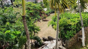 Un jardin luxuriant, avec des palmiers, un sentier en pierre et une vue sur des bâtiments en arrière-plan.