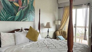 Une chambre avec un grand lit, un tableau représentant des plantes tropicales, une tête de lit en bois, une table de chevet avec une lampe et une vue sur l’extérieur par une fenêtre.
