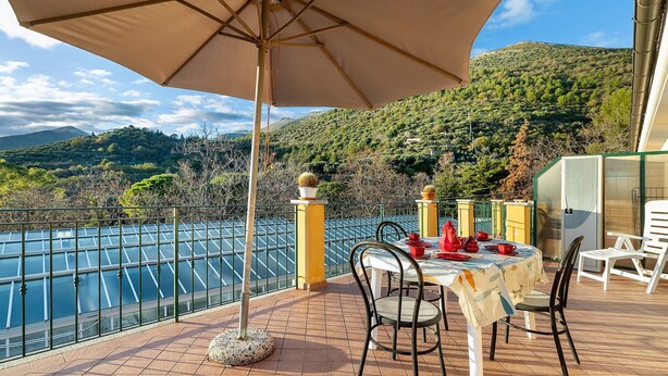 A terrace with a table, chairs, and an umbrella, overlooking a body of water and hills. - ‘Acacia’ Apartment with Sea View, Wi-Fi and Air Conditioning (Ceriale)