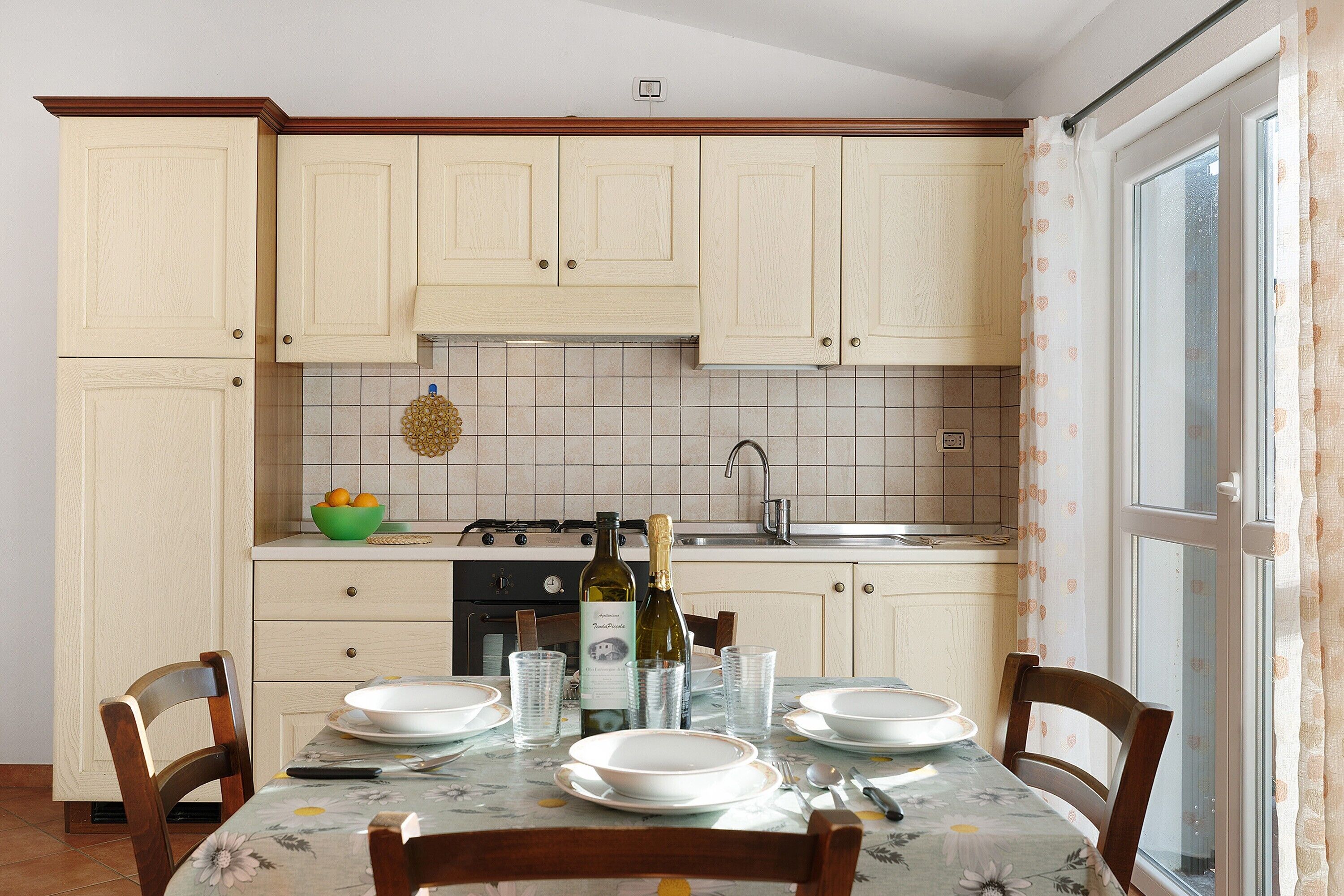 Una cucina con mobili chiari, un tavolo da pranzo apparecchiato con piatti e bicchieri, e una bottiglia di vino.