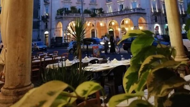 An outdoor dining area with tables and chairs, a building with multiple balconies, and an arched structure with a hanging light fixture. - B&B Piazza Mazzini (Catania)