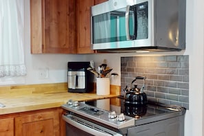 A kitchen with a stainless steel microwave, a kettle, a coffee maker, and wooden cabinets.
