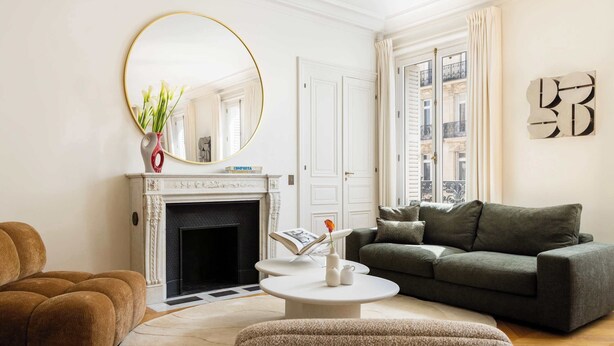 Un salon avec une cheminée, un miroir rond, un canapé vert et une table basse ronde.
