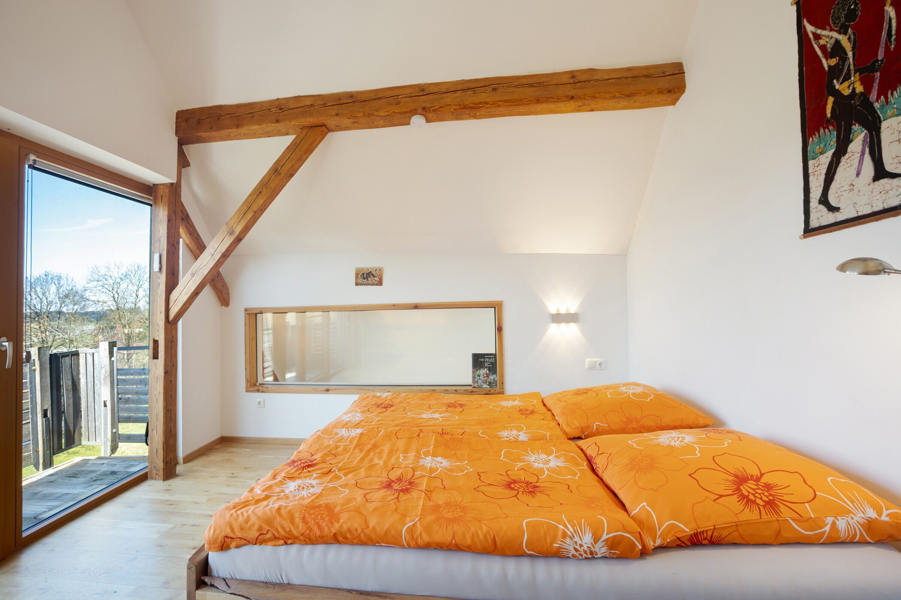 Une chambre avec une poutre en bois au plafond, un lit recouvert d’une couette à motifs floraux orange, une grande fenêtre et un miroir fixé au mur.