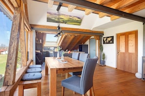 A dining area with a wooden table and chairs, a view of the outdoors, and a ceiling with exposed beams.