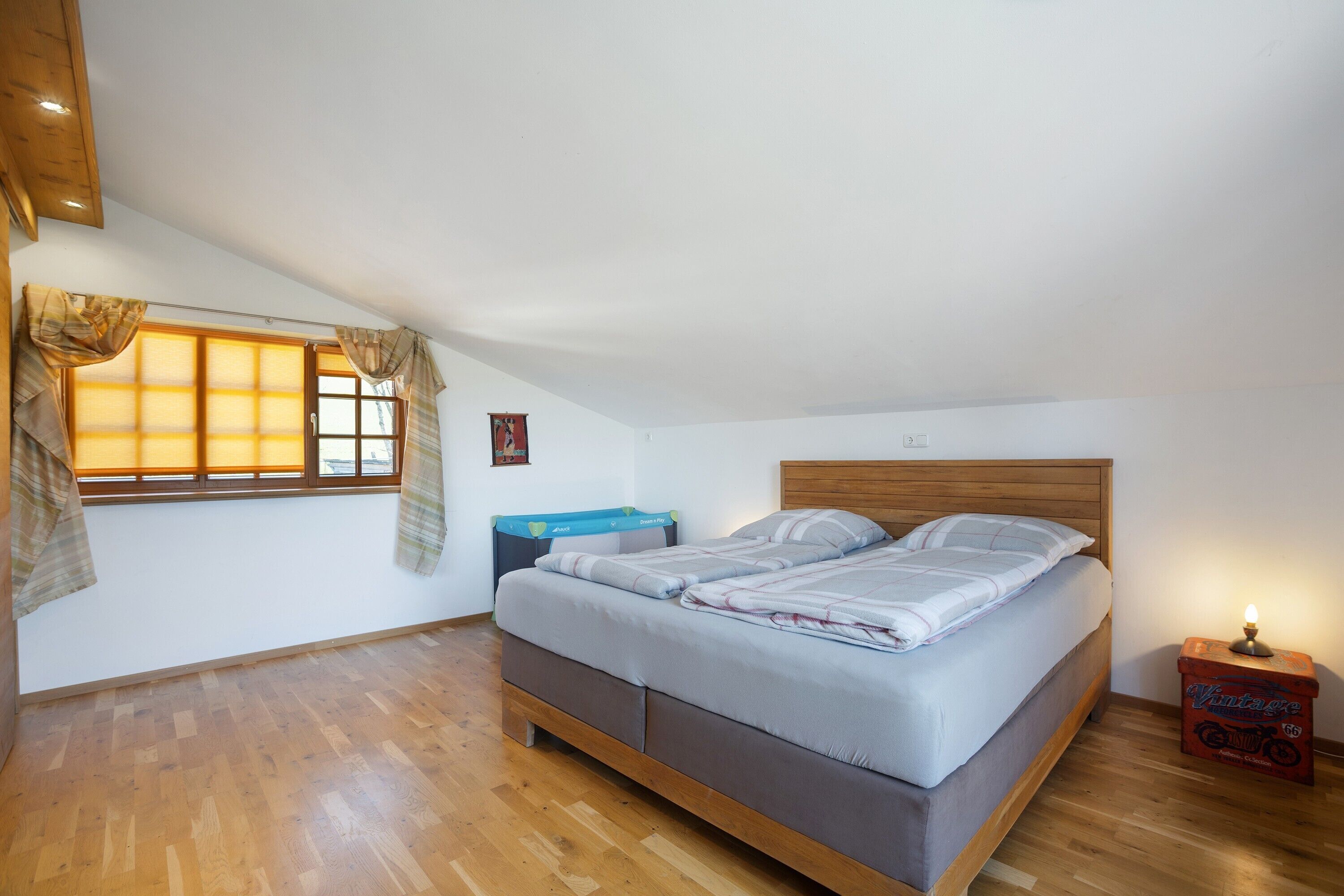 Une chambre à coucher avec un lit, un plancher en bois, une fenêtre avec des rideaux, une table de chevet et une petite table de nuit avec une lampe.